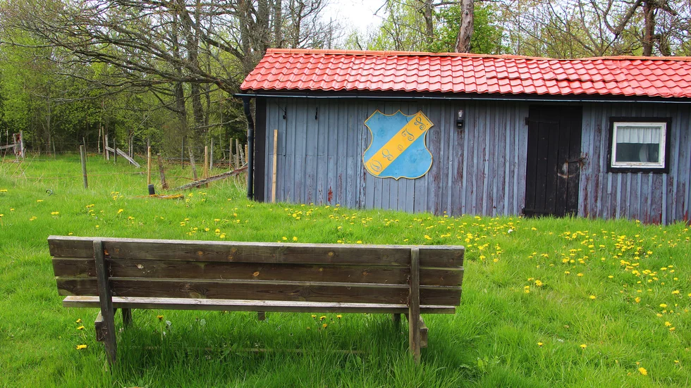 På Örtomtavallen kan man, om så önskas, vända ryggen åt planen och istället granska den gamla klubbstugan med det ursprungliga klubbmärket på väggen.