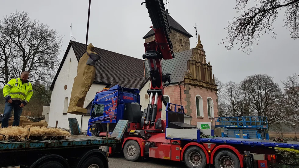 En av de stora skulpturerna lastas av. I bakgrunden syns Vallstanäskoret som är i rött tegel, till skillnad från resten av kyrkan.