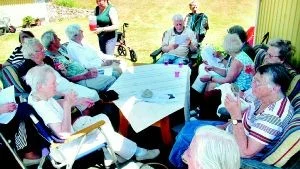 Grillfest. Gästerna har bänkat sig runt bordet hemma hos Inger Johansson på Tättö. Längst bak på bildens ses Röda Korsets ordförande Ursula Fernström servera förfriskningar och bredvid henne ses Lilian Lijedahl med sitt dragspel.