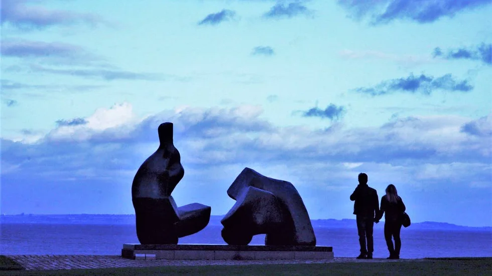 Två besökare bredvid den stora skulpturen av Henry Moore medan skymningen sänker sig över Öresund.