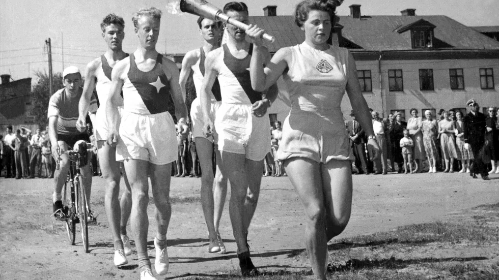 OS-facklan kommer till Luleå den 9 juli 1952. Många har samlats för att få en skymt av den olympiska facklan som bärs av skidåkaren Sonja Edström. Bakom syns Alf Sandström på cykel, friidrottarna Stig Olsson, P O Öhman, Ingemar Nilsson och Holger Nyman. De olympiska spelen avgörs i Helsingfors.