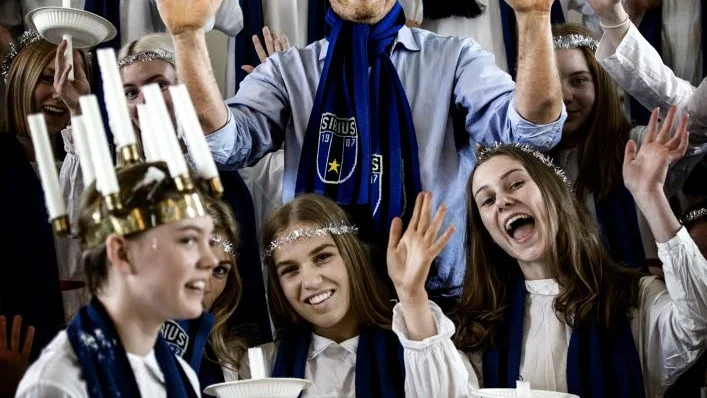 Luciatåg. Jacke Sjödin gör succé med sina snälla Siriusfilmer där motståndarlagen välkomnas till Uppsala. Här står han – mitt i sommaren – omgiven av Uppsala Musikklassers luciakör, som stod för skönsången i den senaste filmen.