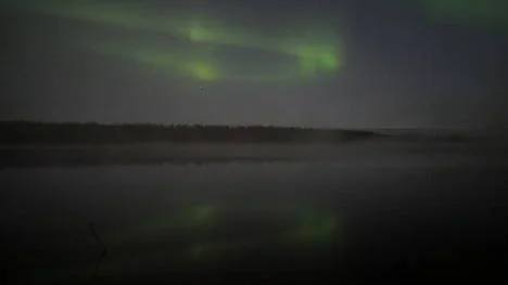 En klar och stilla natt där dimman sakta rullar in över vattnet och norrskenet som dansar över himmelen speglas i vattnet