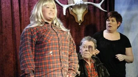 NYKOMLING I JAKTLAGET. Anne-Lie Eriksson är jaktlagets nya Baby Doll, som just har märkt Roland Christoffersson med en blåklocka, allt medan Ingela Johansson förekommer med öppen mun och den tidigare nedlagda älgen betraktar alltsammans från sin plats på väggen.