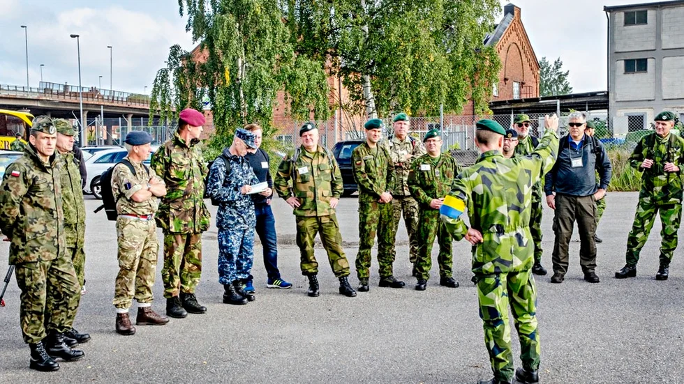 En buss med cirka 15 utländska observatörer har precis anlänt utanför hamnens grindar och tittar på när hemvärnssoldaterna strider mot fienden ”A-land”.