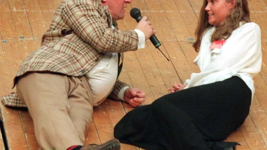 Kurt Olsson (Lasse Brandeby), i avslappnad golvpose, intervjuar luciakandidaten Karin Engman på höstmässan 1995. 