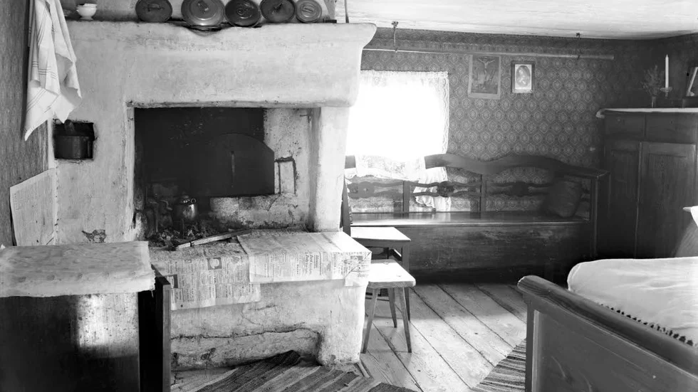 Interiör från ryggåsstugan på Djurgårdsgatan 3 år 1903. Bostadens enda rum fungerade som kök, vardags- och sovrum. I spisen står en kaffekanna framför luckan till bakugnen. Invid fönstret står en dragsoffa. På väggen hänger två tryck, det ena föreställande den korsfäste Kristus, den andra Jungfru Maria. Till vänster ses kommoden med handdukshängaren ovanför. Till höger skymtar den uppbäddade sängen. Utdragna kunde soffan och sängen fungera som sovplats för ganska många.