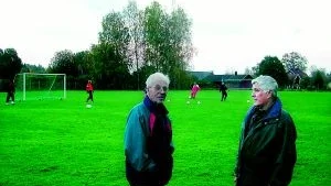 Torvald Andersson och Gösta Carlsson vid Torvalla fotbollsplan. Båda har ägnat massor med tid och energi åt sin förening, både som aktiva idrottsmän och genom ideella arbetsinsatser. Torvald är också den som fått ge fotbollsplanen dess namn. I bakgrunden tränar slocknade stjärnor inför lördagens drabbning.