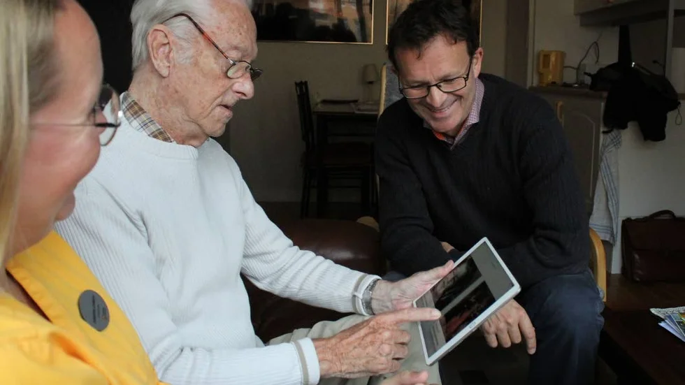 Hemma hos Birger. Josehine Lindblom från Linköpings kommun, Birger Rönnbäck och sonen John tittar på gamla bilder i appen Levnadsberättelser från Linköpingsföretaget Identive.