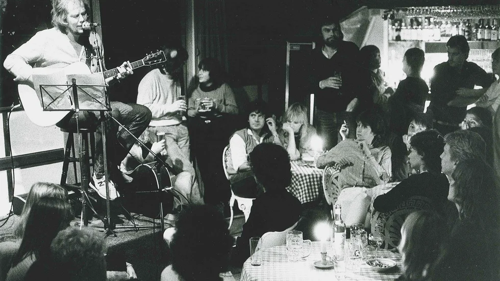 Ola Magnell var flitig på Gotland med sin gitarr under 70- och 80-talen, ofta ordnades spelningarna av nära vännen André Lahovary. Det är han som sitter i mitten av bilden vid de rutiga bordsduken på den här bilden från 1982.