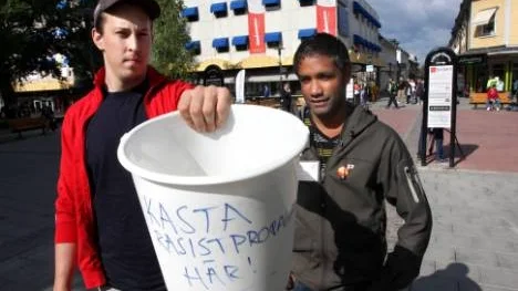 Joel Karlsson, Ung vänster, och Fredrik Hansson, SSU, med en hink där människor uppmanas kasta Sverigedemokraternas material.