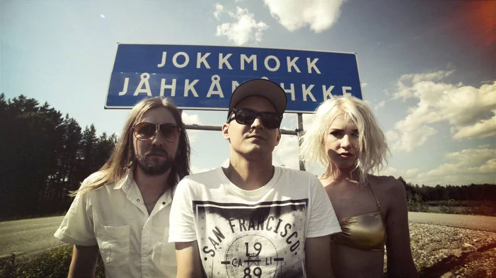DÄR VÄGARNA VÄNDER. Jokkmokkssonen Magnus Ekelund, alias Kitok, vid Jokkmokk/Jåhkåmåkkeskylten, flankerad av Erik Zettervall och Stina Vikström.     Bilden är tagen från den genomsnygga videon till Paradise Jokkmokk.