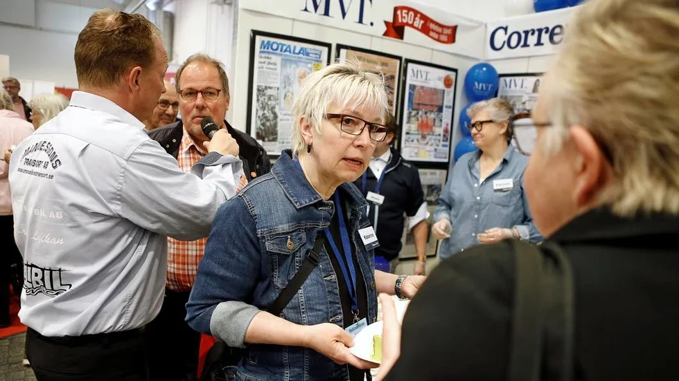 Vadstenareporter Katarina Hydén, för dagen tårtgeneral, serverar tårta i MVT/Correns monter. I bakgrunden intervjuas Johan Andersson (S).