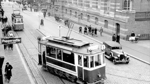 Livfull vy från 30-talets början. Vänstertrafik gäller, båda spårvagnarna är således på väg nerför Drottninggatan. Tvåan från Kneippbaden kör mot  Marielund. Medan ringlinjen, fullbordad 1926, gjorde skäl för namnet där den gick innerstaden runt, runt. Omedelbart till vänster Holmströms & Rockströms pappershandel, följd av Rosanders herrekipering. Längre bort skymtar det stora hotellet, som dåförtiden ännu inte hette Grand utan fortfarande Göta. Fotografiet tillhör Stadsarkivet.