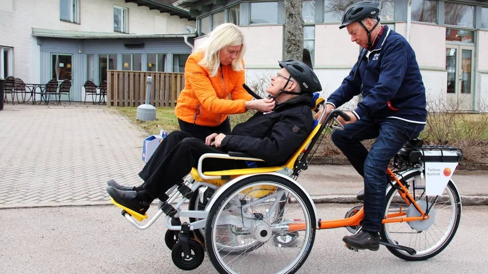 K-G Holmsten testcyklar och Karl-Erik Engdahl testar rollen som passagerare, men Suzanne Gehlin från Vadstena Sparbank vill att de ska ha hjälm så att de åker säkert.