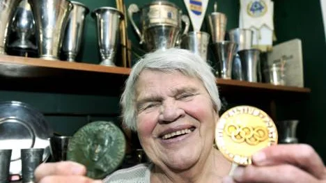 Sonja Edström-Ruthström visar upp sina olympiska medaljer. Bakom hennes syns den välfyllda prishyllan.