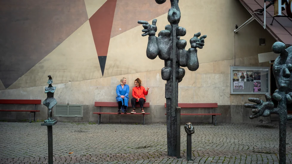 Bror Marklunds verk "Gycklarna" pryder torget i Årsta där repetitionerna av Norrbottensteaterns "Köp Hjärtan" påbörjades. På bilden tar Vanna Rosenberg och Karin Paulin en paus med utsikt över gycklarsällskapet i brons.