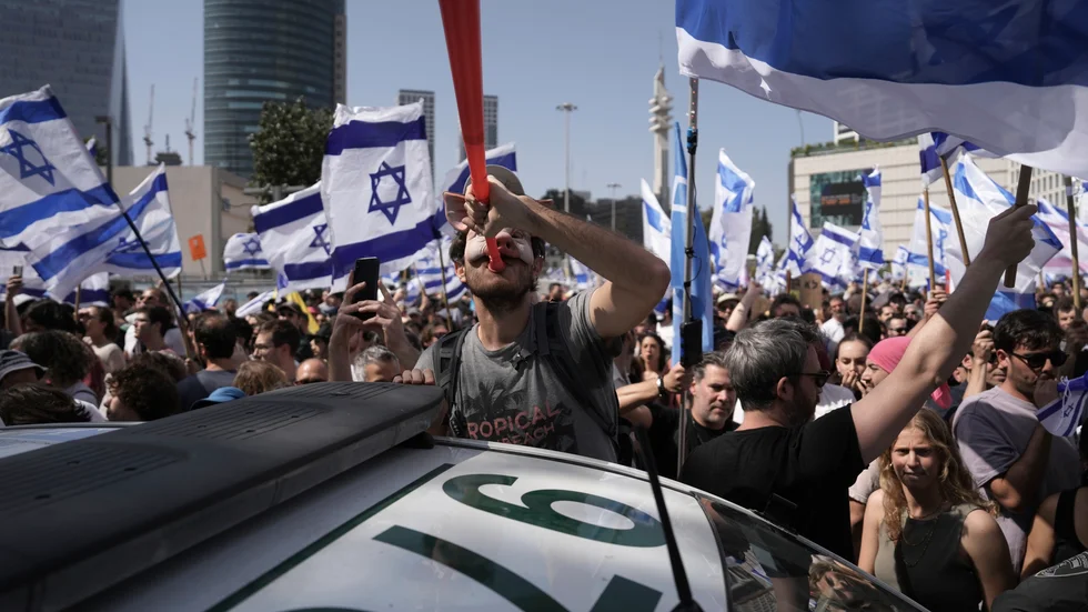 En demonstrant i grismask blåser i en vuvuzela bredvid en polisbil i Tel Aviv.
