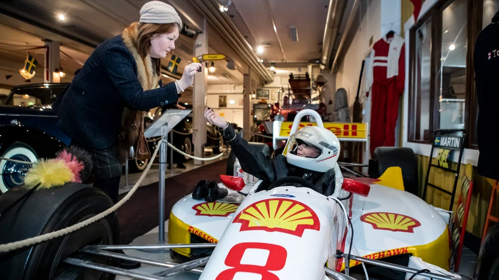 Konsulinnan Lobb (Lotta Karlsson) räcker över en nyckel till Kenny Bräck (Linnea Nilsson) som sitter i hans bil. De är två av de karaktärer deltagarna får möta i sportlovsguidningarna på Motala Motormuseum.
