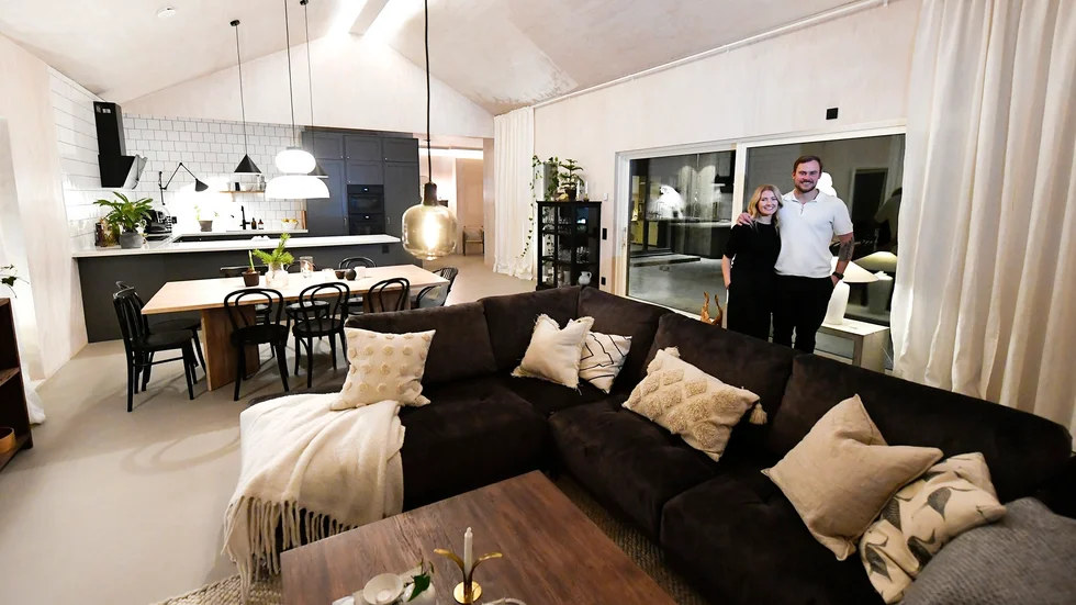 Lydia Broström och Alexander Brinkmann står i vardagsrummet. På väggar och tak syns plywoodskivorna och på golvet den ljusgrå, matta betongen som har flytspackel ovanpå. 
