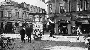 Liv och rörelse i utkanten av Nya Torget, någon gång under år 1938. Det är Hospitalsgatan som försvinner bakåt in i bilden, och Kristinagatan (Östra Kyrkogatan dåförtiden) som drar från vänster till höger genom bilden. I korsningen låg Sterns klädaffär, så välbekant för äldre Norrköpingsbor. Invid entrén till hörnhuset till vänster berättar en skylt om Konsum Specerier. Och rakt fram i bilden: Skomagasinet.
Fotografiet tillhör byggnadsnämndens samlingar i Stadsarkivet.