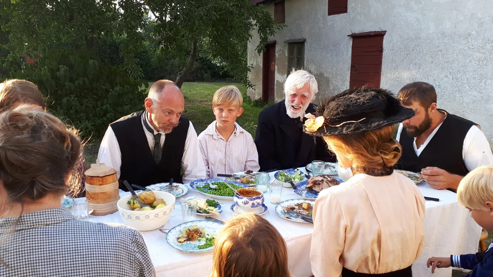 Samlade till den årliga sommarmiddagen. Anna (Julia Westberg) i hatt med ryggen mot kameran hyser en längtan som inte ryms i de planer vitskäggige svärfadern (Göthe Carlsson) har för framtiden. 